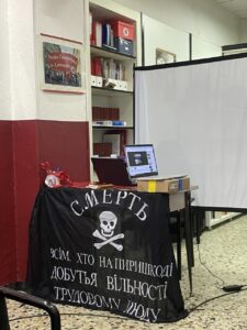 Un tavolo con un computer portatile e dei fogli è coperto da una bandiera nera dell'esercito di Makhno con un teschio e ossa incrociate, accanto a uno schermo di proiezione in una stanza con scaffali e un manifesto del Partito Comunista.