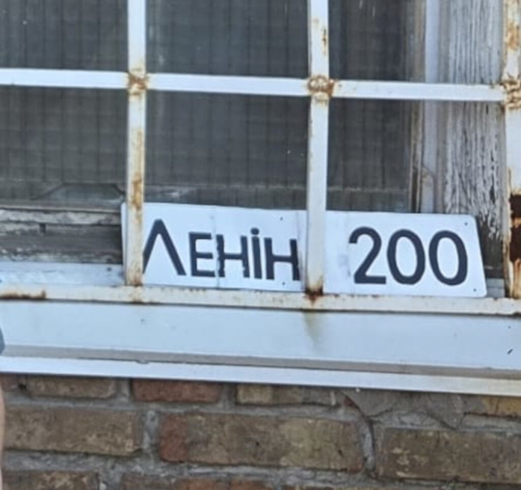 Primo piano di una finestra usurata con una grata metallica arrugginita, dietro la quale si vede un cartello con scritto “ЛЕНІН 200” (Lenin 200).